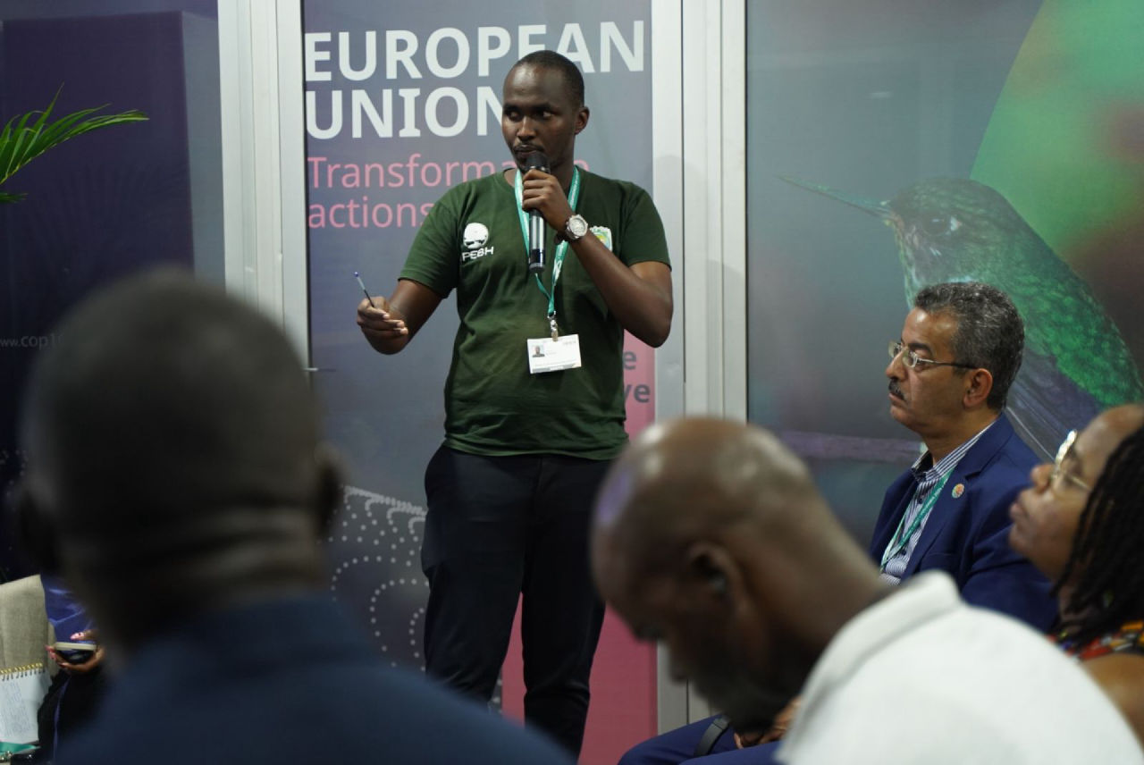 Un représentant de l’APEBH prend la parole avec un microphone devant le visuel du Pavillon de l’Union européenne.
